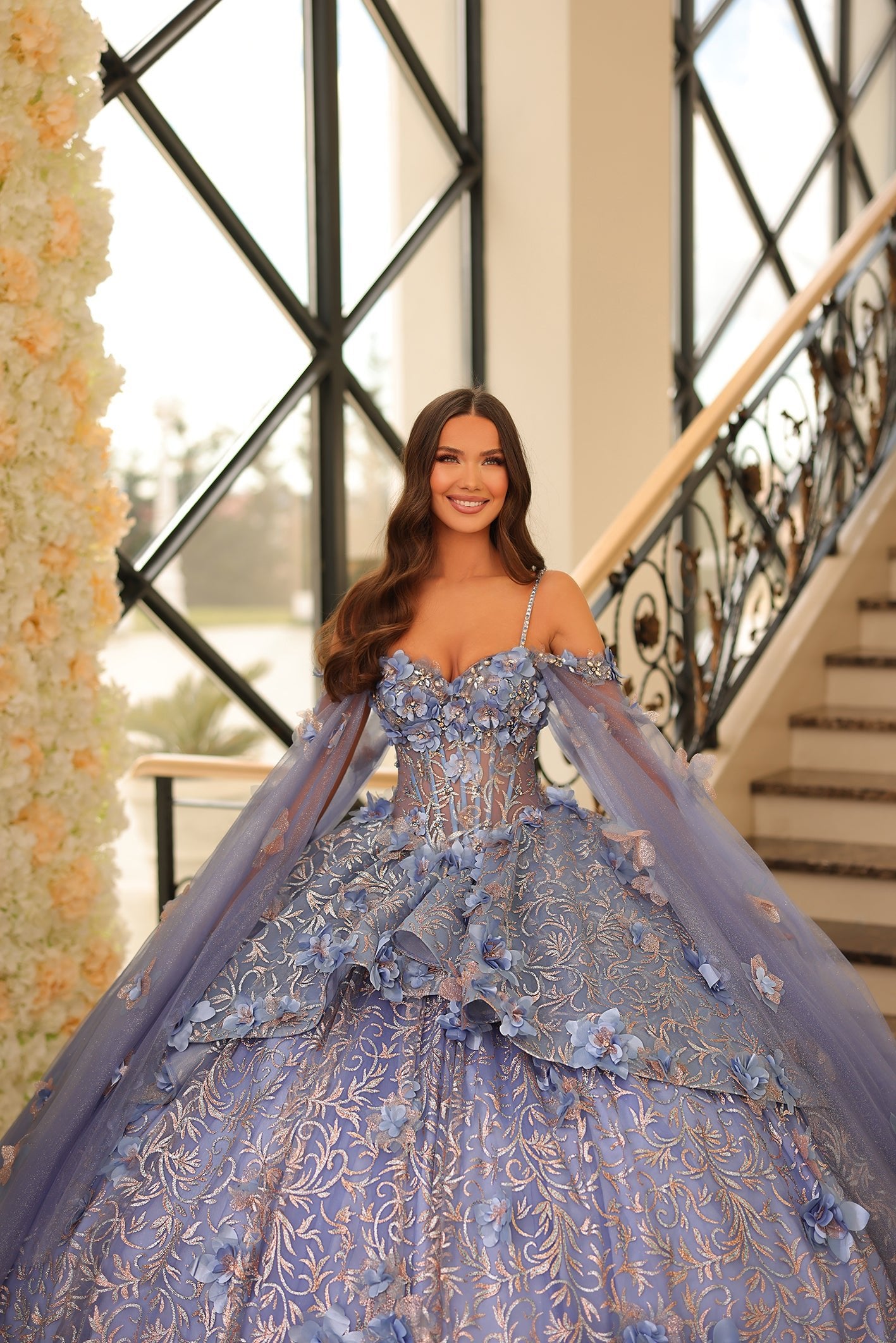 A woman wears a lavender blue ball gown with 3D floral details, standing on an elegant staircase with ornate windows.