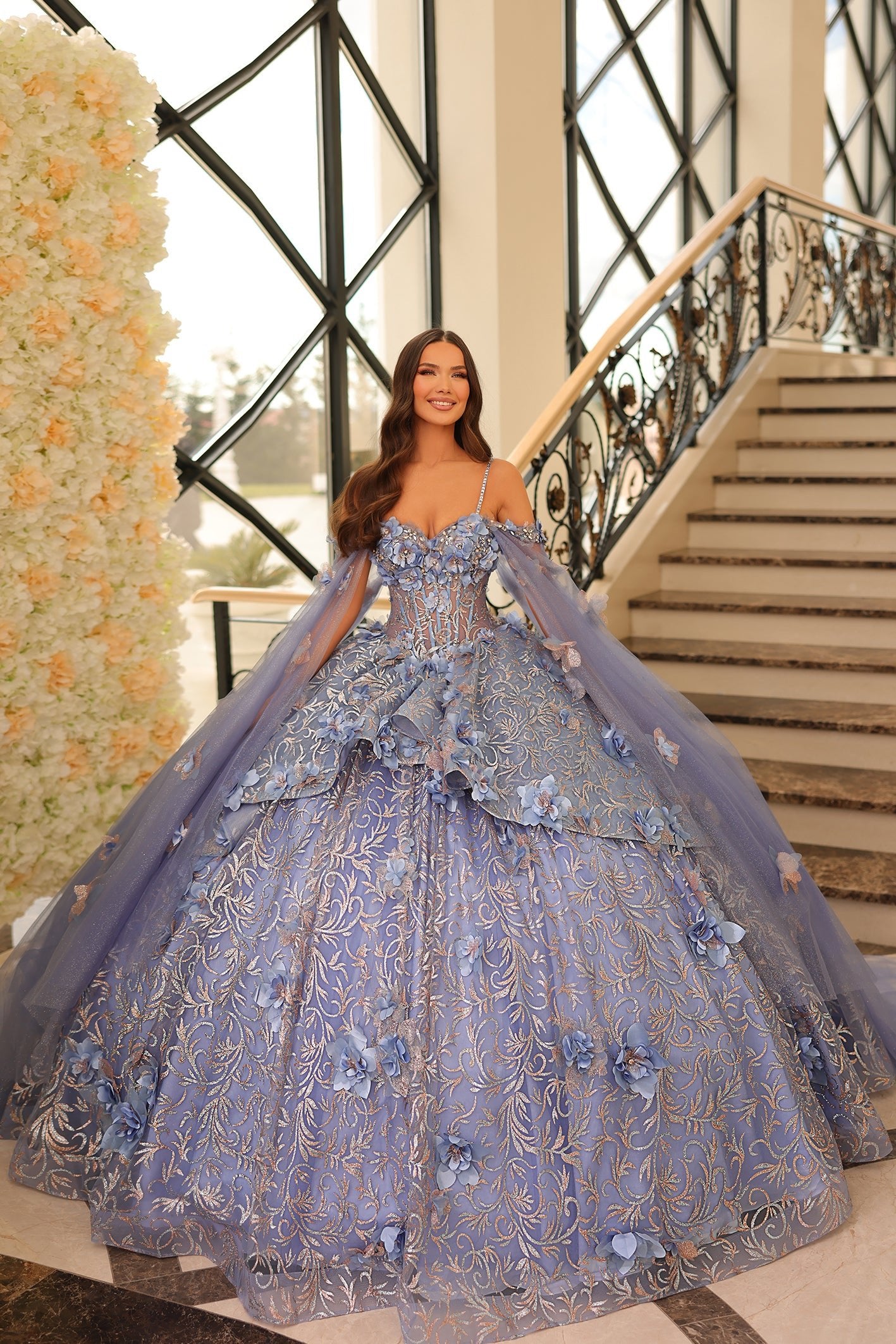 A woman wears a voluminous Dusty Blue ball gown with floral appliqués and delicate straps near an ornate staircase.