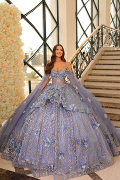 A woman wears a voluminous Dusty Blue ball gown with floral appliqués and delicate straps near an ornate staircase.