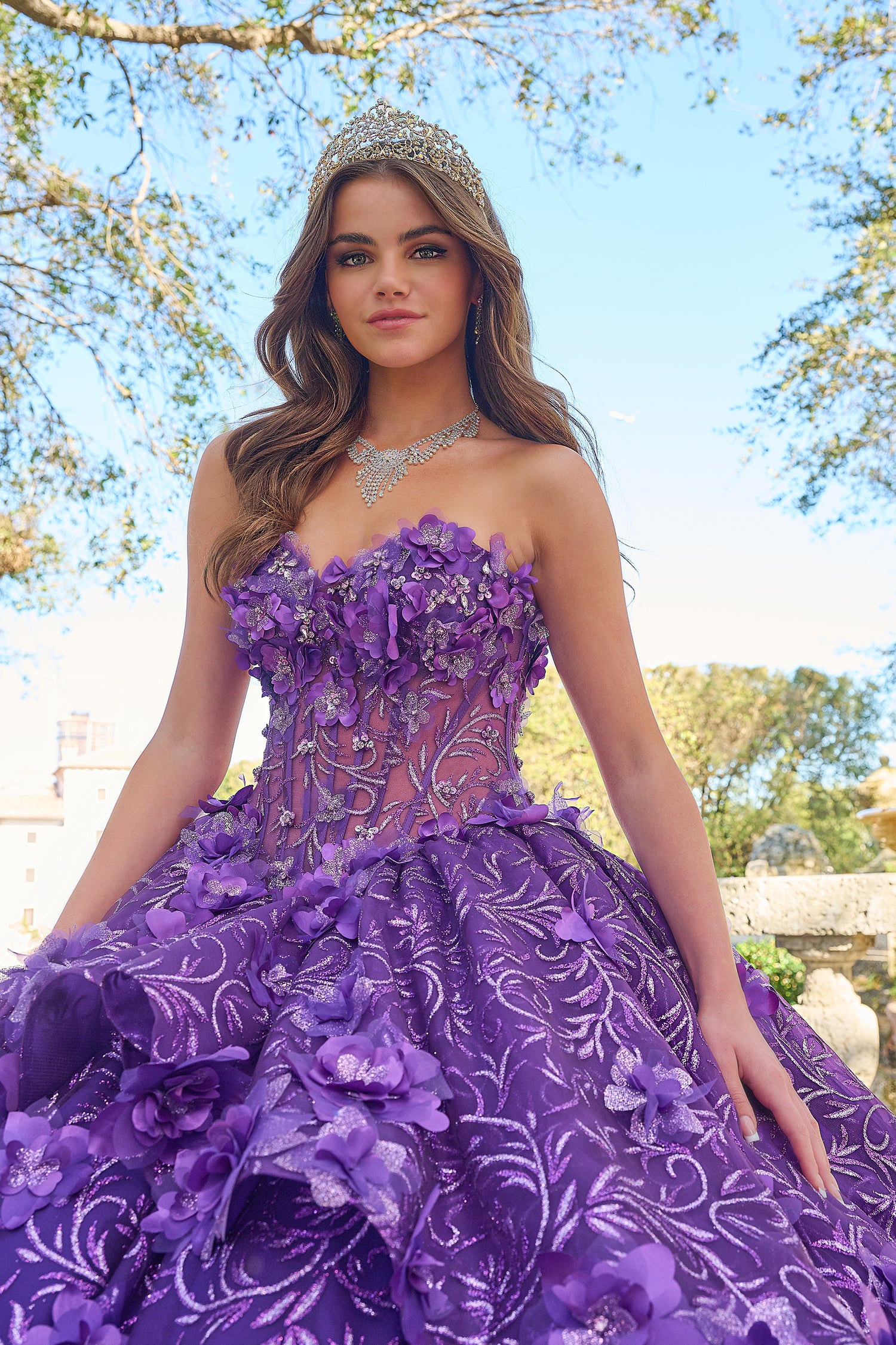 A young woman wears a purple floral ball gown with a tiara and sparkling necklace in an outdoor setting.