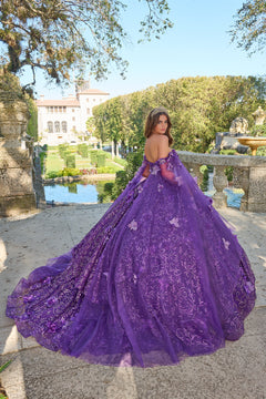 A woman in a voluminous purple off-shoulder ball gown poses in a garden with a historic mansion in the background.