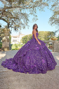 A woman wears a voluminous purple floral quinceañera gown in a garden with stone pillars and trees.