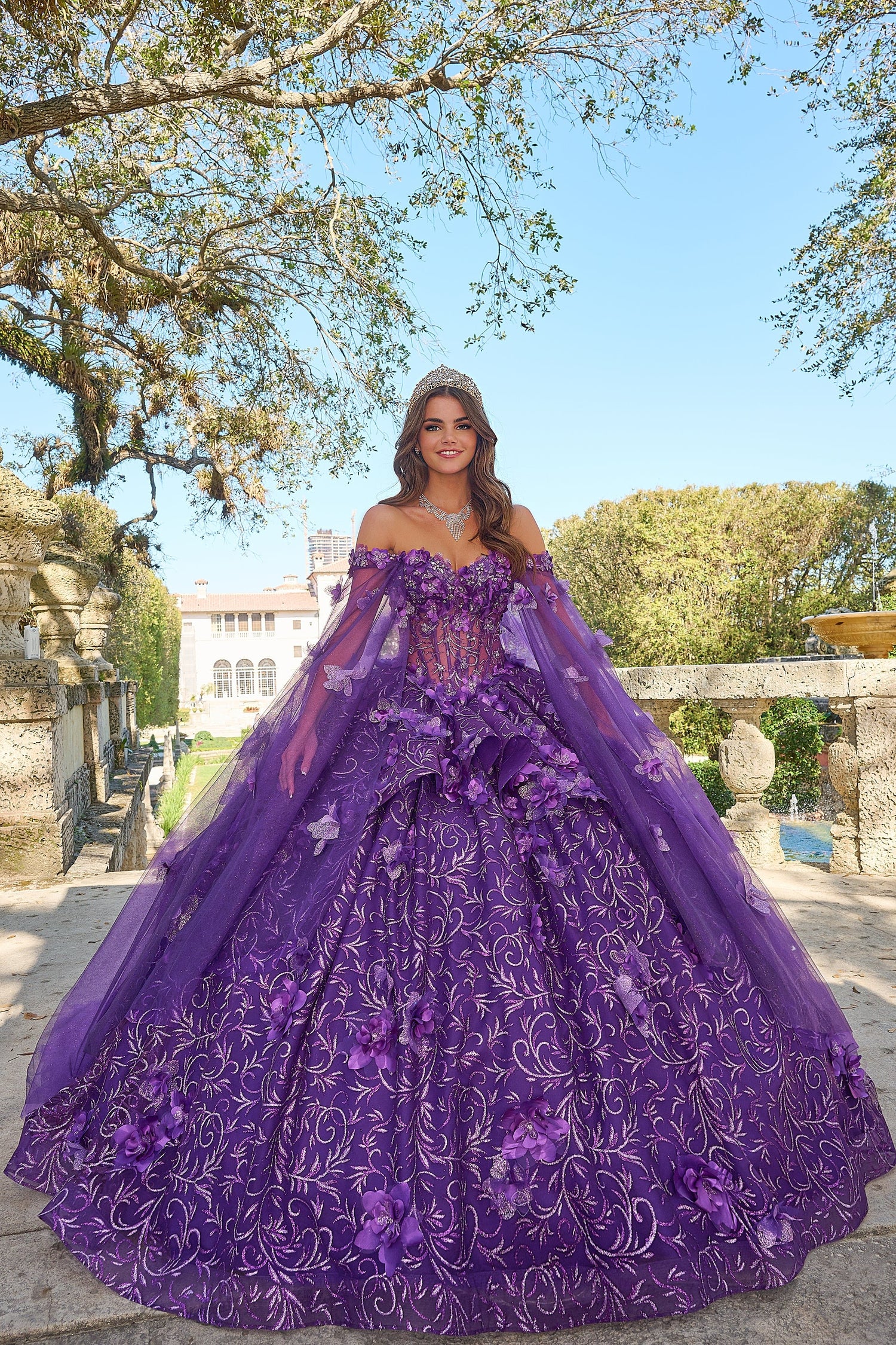 A woman wears a lavish Purple ball gown with 3D floral details and a tiara in a garden setting.
