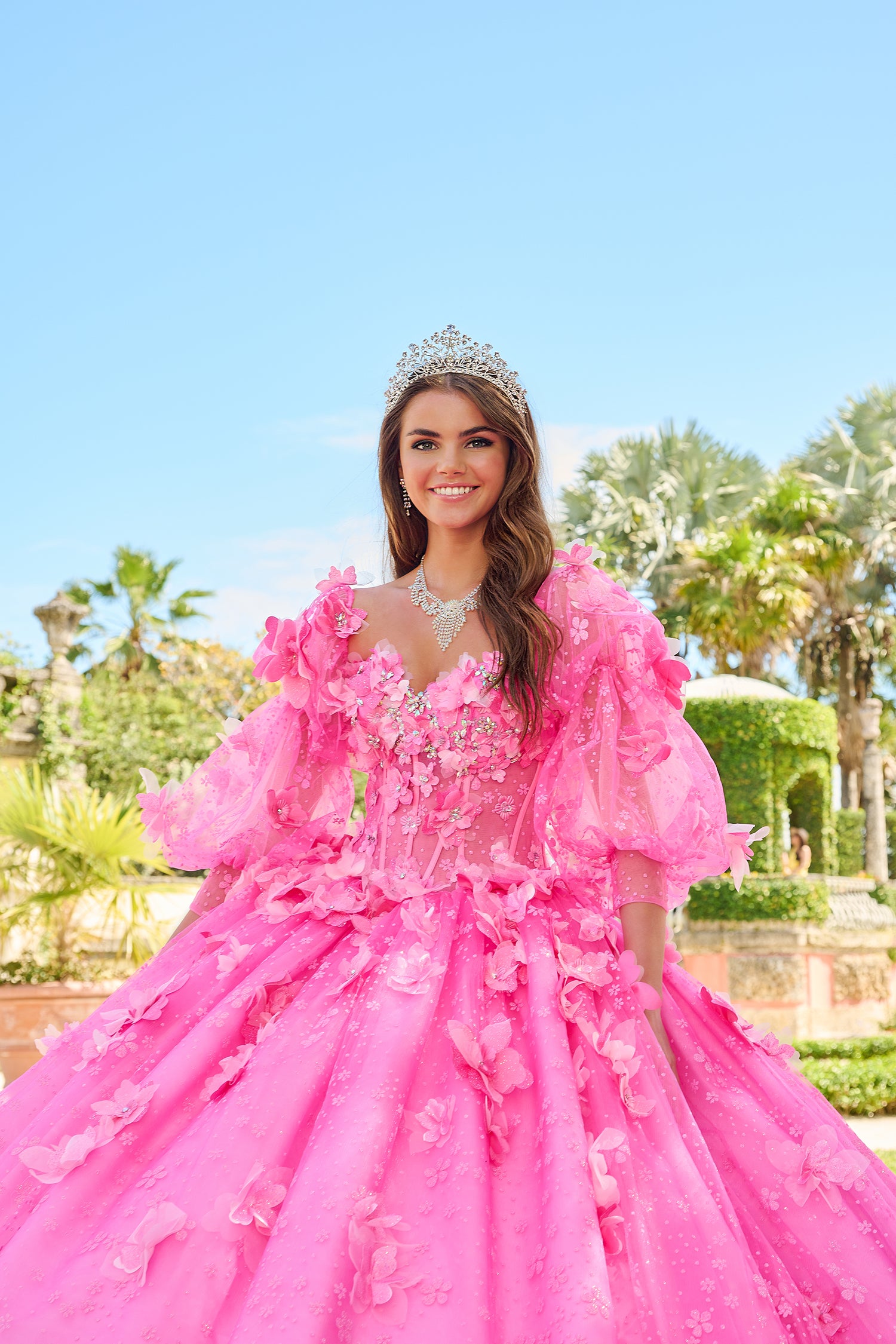 A smiling young woman wears a voluminous pink floral gown and tiara in a garden setting.