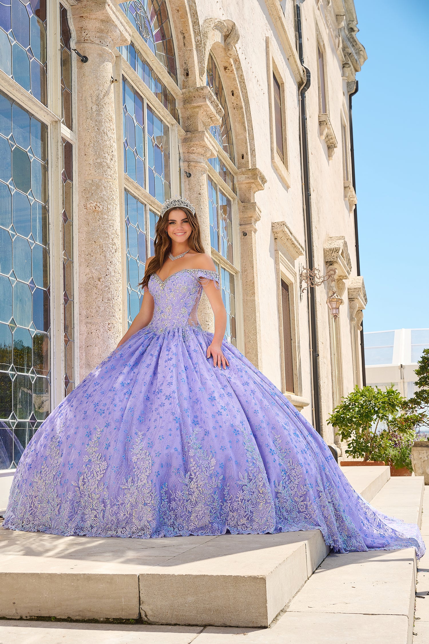 A woman in a lilac ballgown with intricate lace details stands on steps near ornate windows in a sunlit setting.