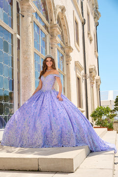 A woman in a lilac ballgown with intricate lace details stands on steps near ornate windows in a sunlit setting.
