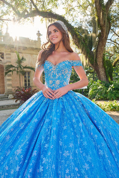 A young woman in a bright blue sparkly quinceañera ball gown with an off-shoulder bodice stands in a lush garden.