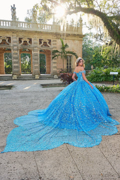 A woman in a voluminous blue lace quinceañera gown poses in a garden with ornate stone architecture.