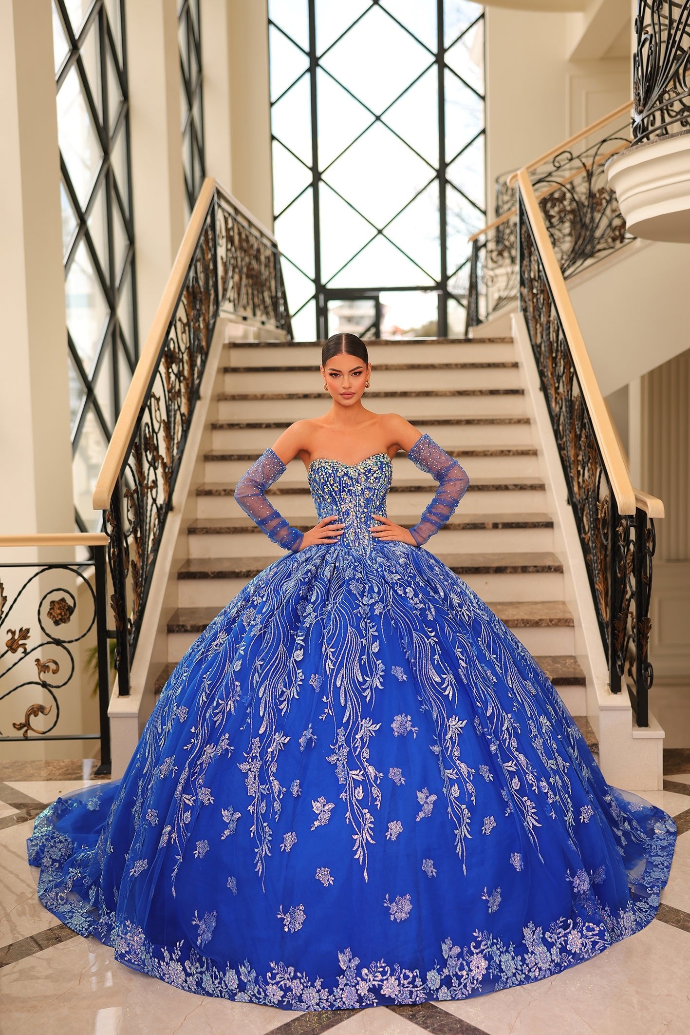 A young woman wears an elaborate Royal Blue ball gown with sparkling embroidery, standing on a grand staircase.
