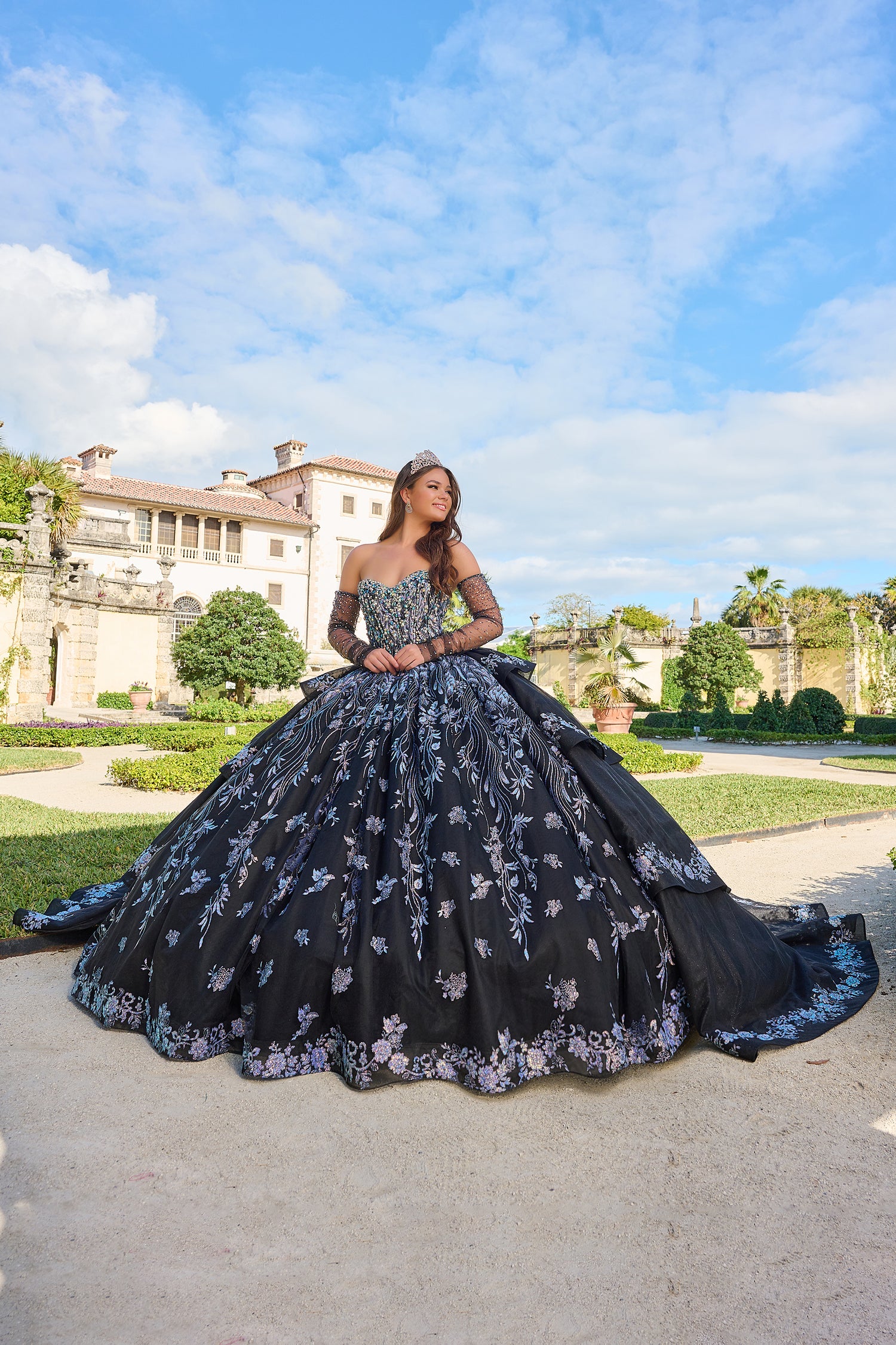 A young woman wears an elaborate black floral quinceañera gown with a tiara, posing in a garden with a historic villa.