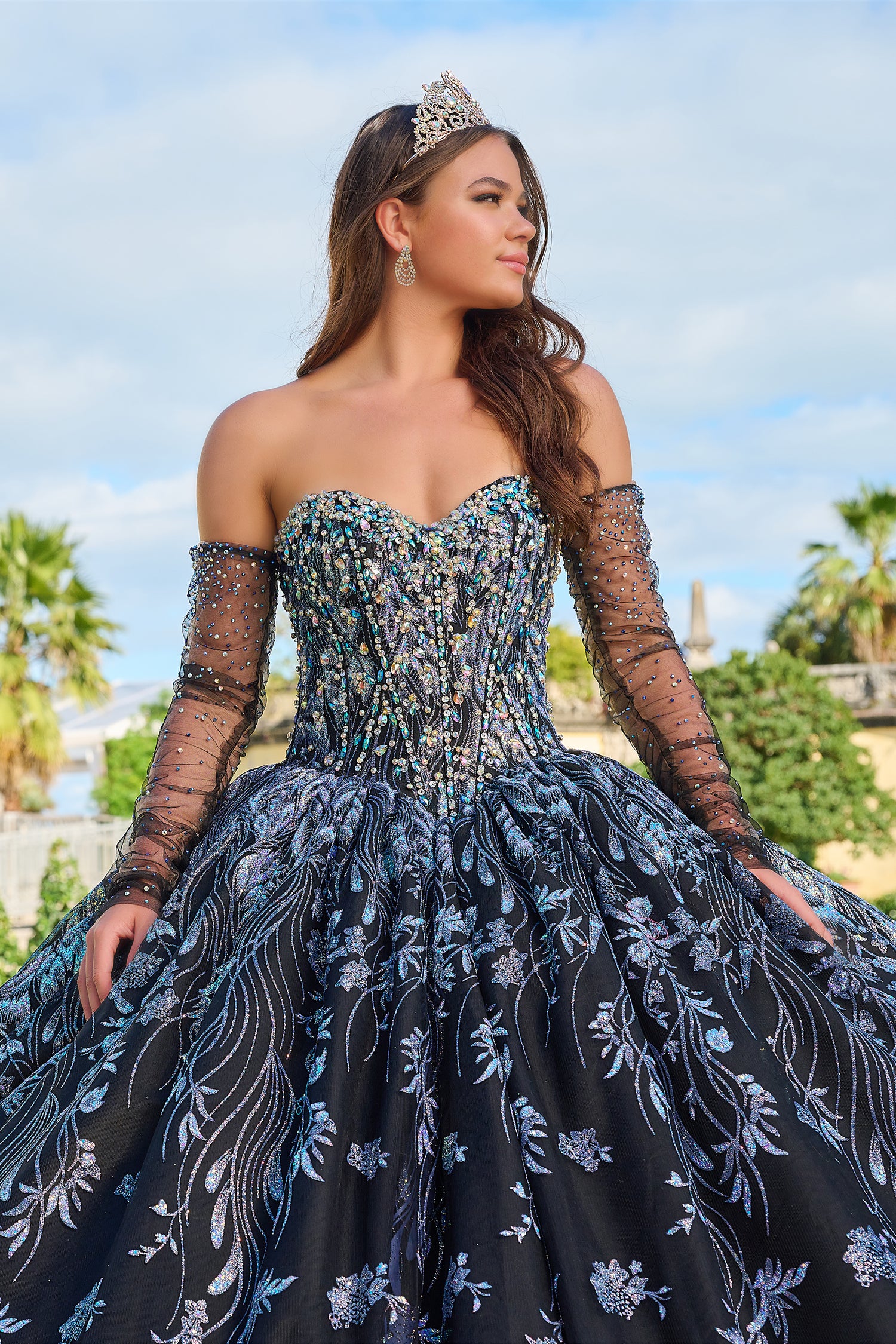 A young woman wears a glittering dark blue quinceañera gown with sheer sleeves and an ornate tiara.