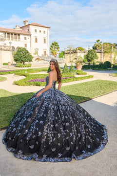 A quinceañera in a black floral gown poses in an elegant garden with a historic villa in the background.