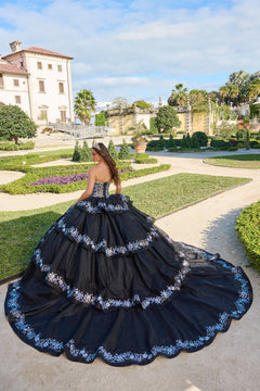 A woman in a black tiered ball gown with blue floral accents poses in an elegant garden with a historic villa.