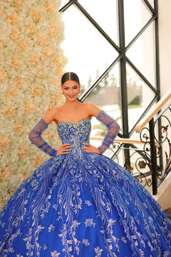 A woman in a blue embellished ball gown poses confidently by a floral wall with ornate window design.