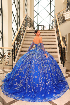 A woman in a royal blue ball gown with intricate floral embroidery poses on an elegant staircase.