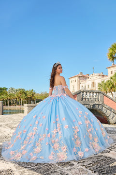 A young woman in a blue floral quinceañera gown poses on a stone bridge with a historic villa in the background.
