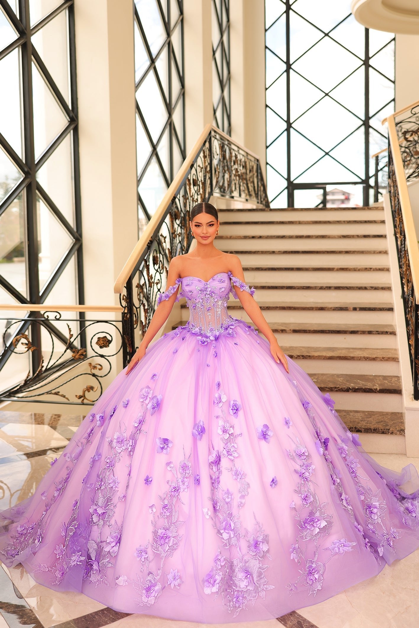 A woman wears a voluminous Lilac/Pink ball gown with 3D floral details, standing on an elegant staircase.