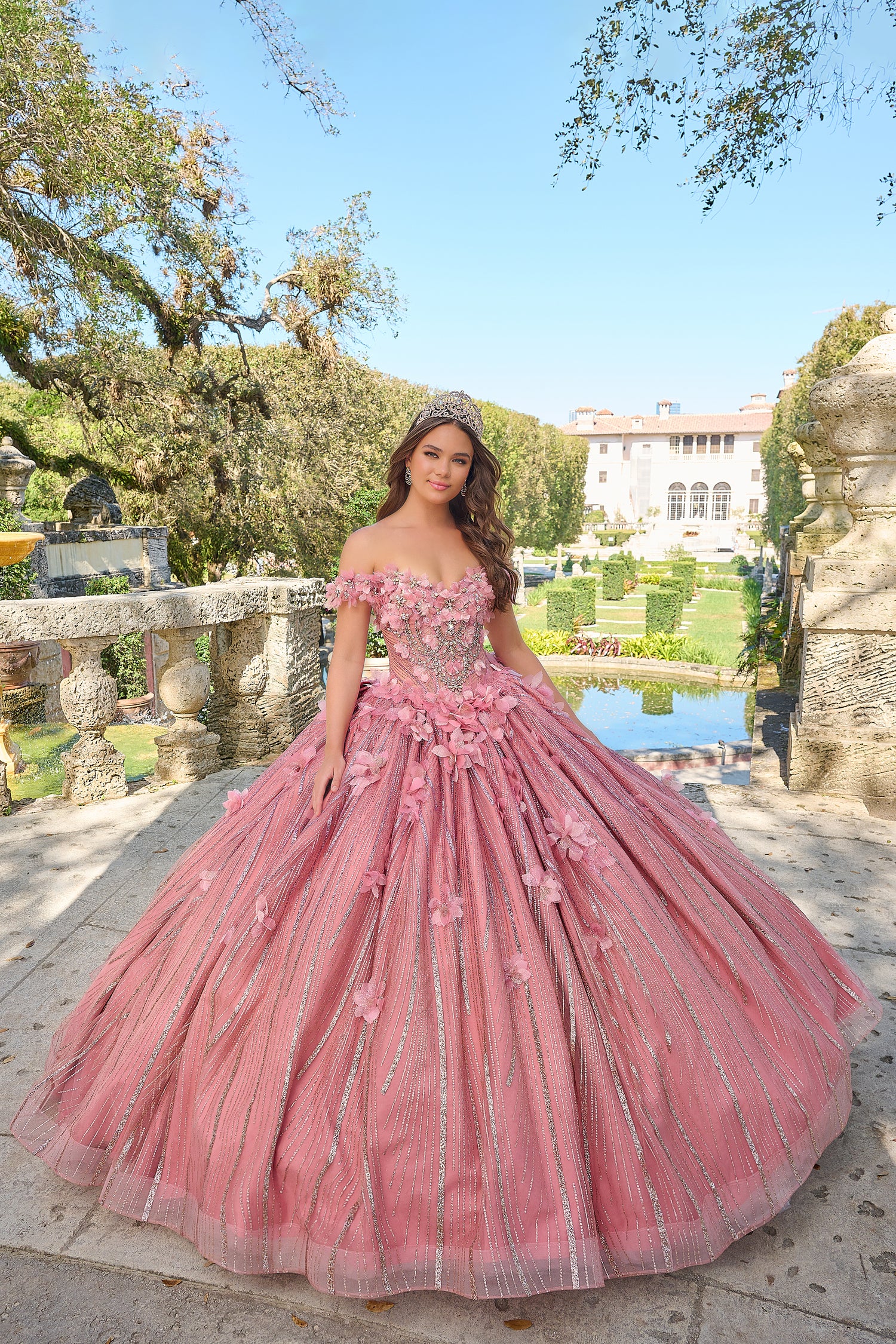 A woman wears a dusty pink off-shoulder ball gown with floral details, standing in a garden with a fountain.