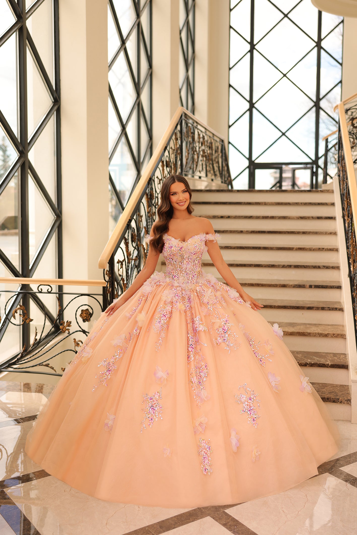 A woman wears a champagne/multi off-shoulder ballgown with floral appliqués, standing on an elegant staircase.
