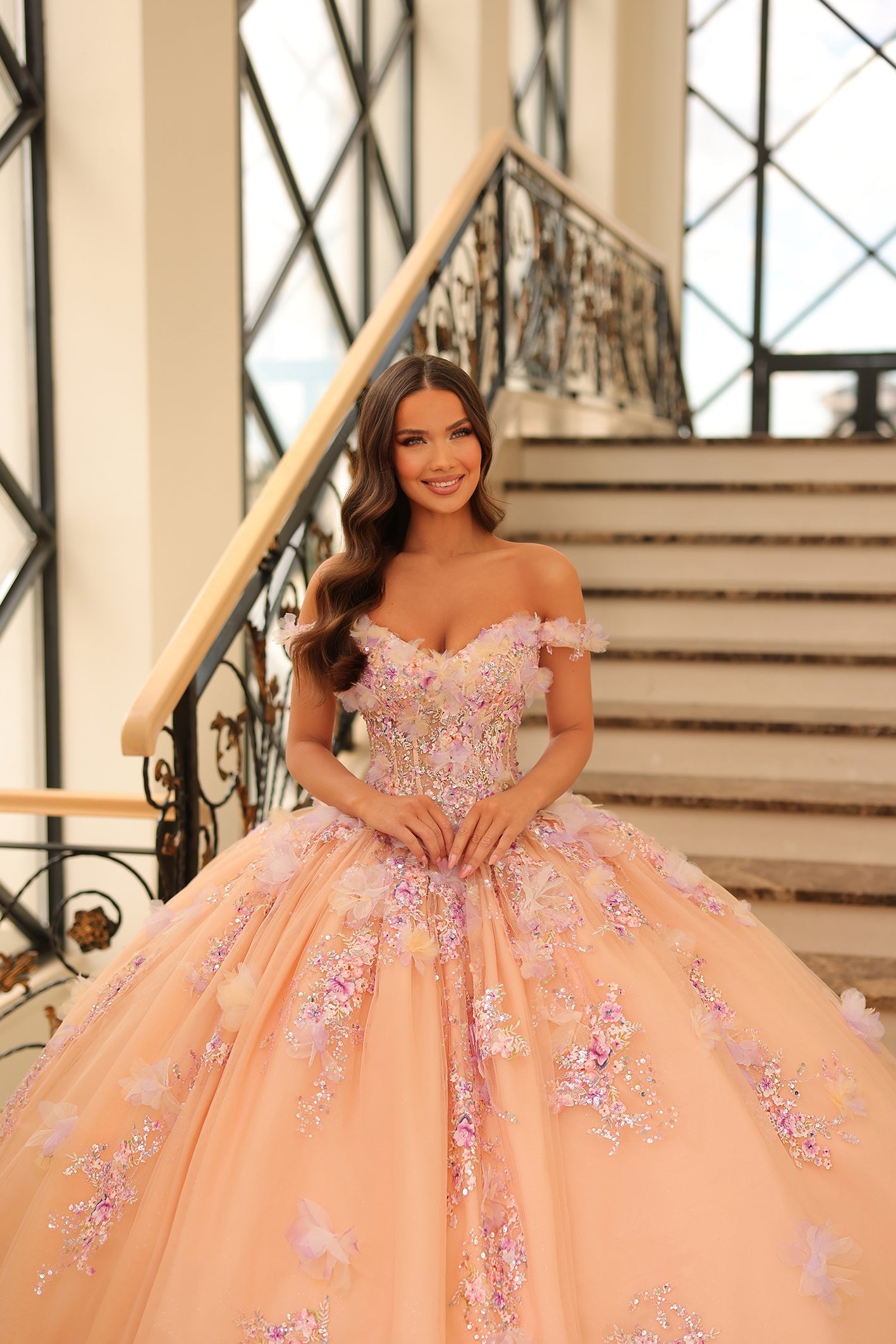 A woman in a pink off-shoulder ballgown with floral embellishments stands on an elegant staircase.
