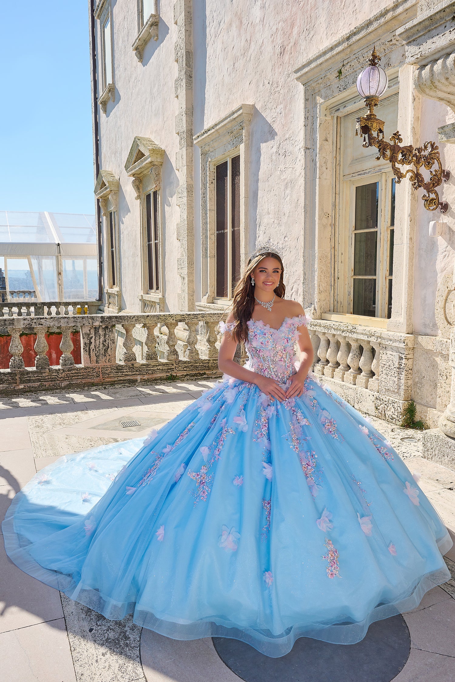 A woman in a Light Blue/Multi floral ball gown stands on a balcony of an ornate white building.