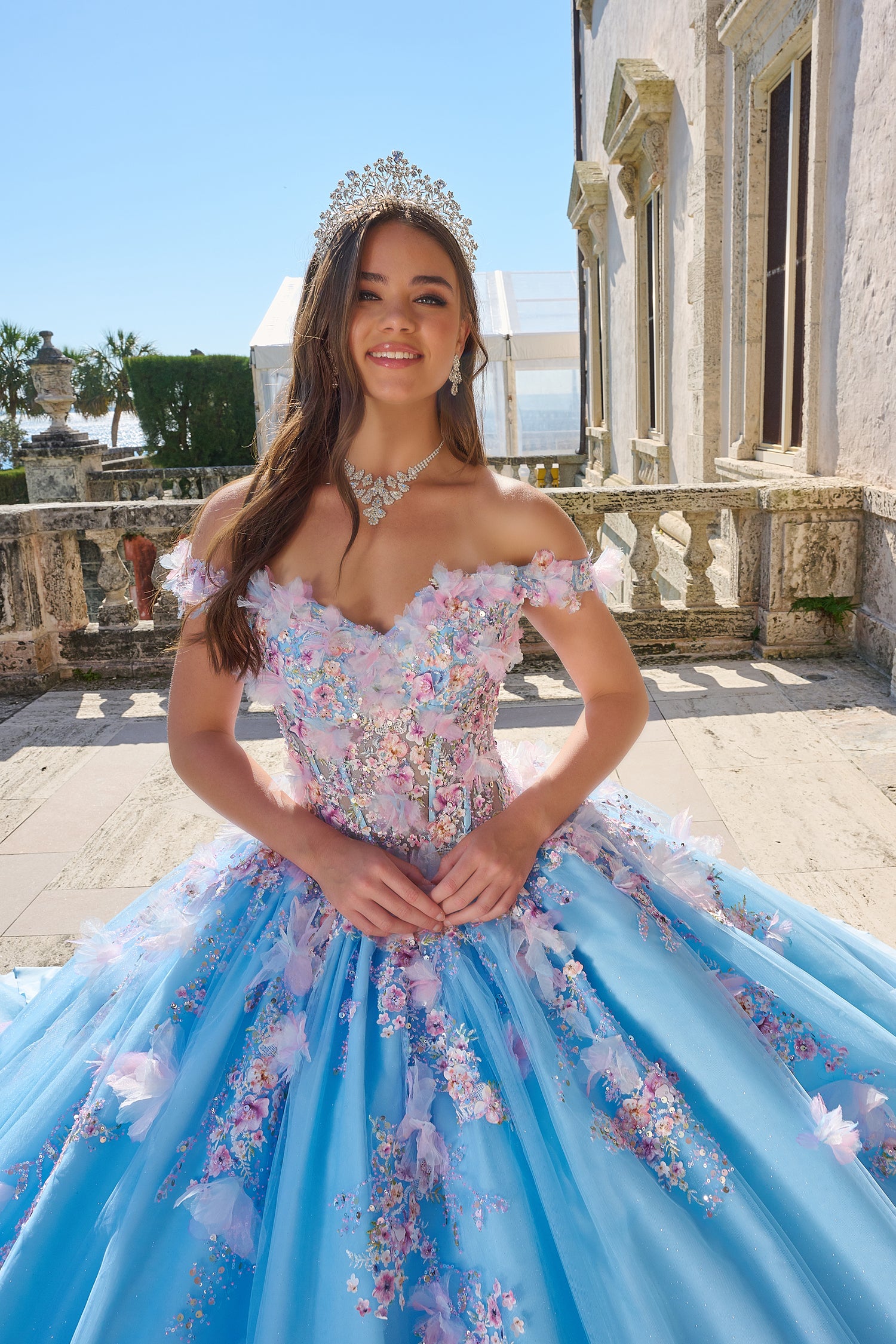 Young woman in a blue and pink floral quinceañera gown with a tiara, standing on a stone terrace.