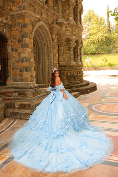 A woman in a sky-blue, off-shoulder quinceañera gown poses before a stone castle with a sweeping, embellished skirt.