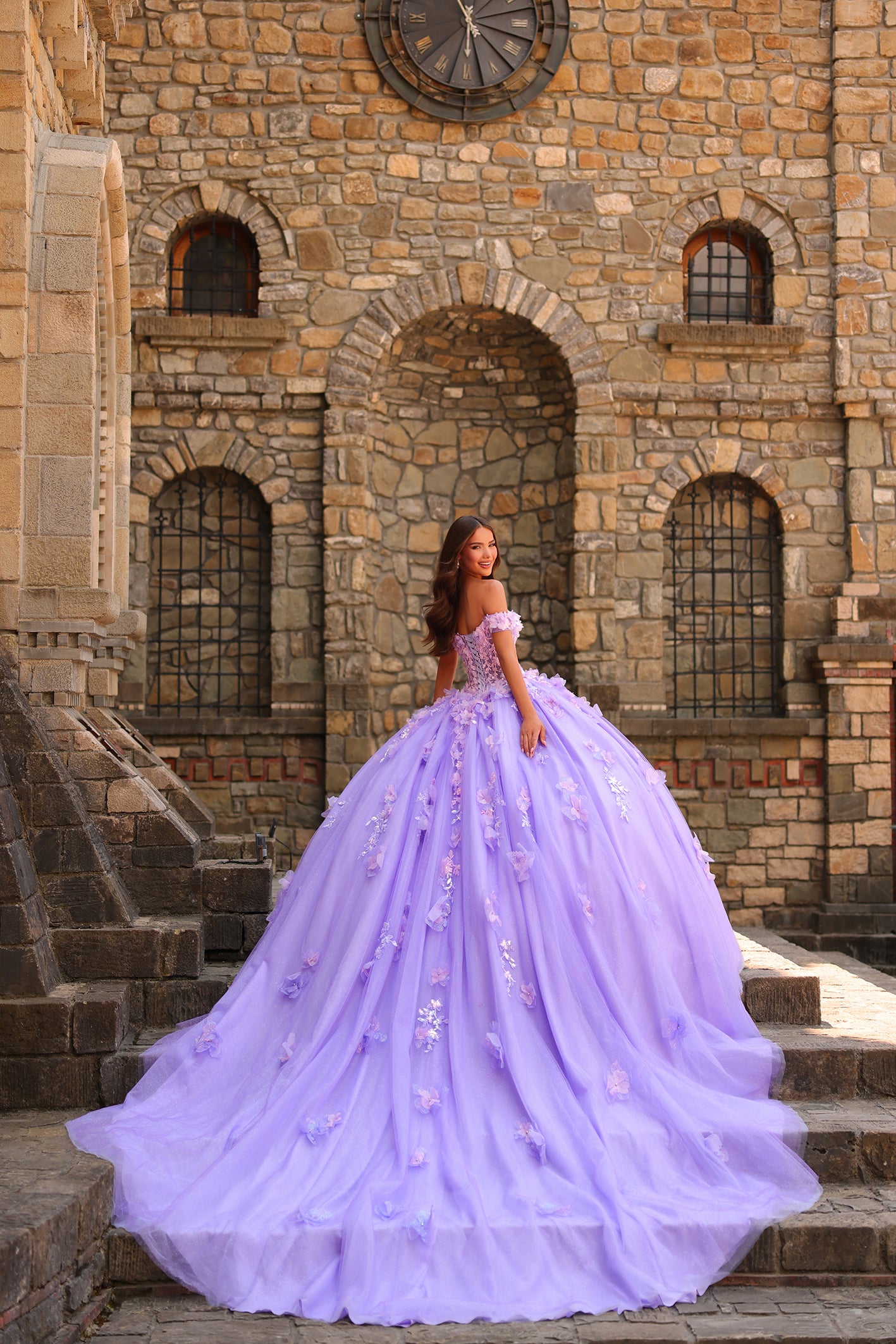 A young woman in a lavender ball gown poses on stone steps before a medieval-style stone building with arched windows.