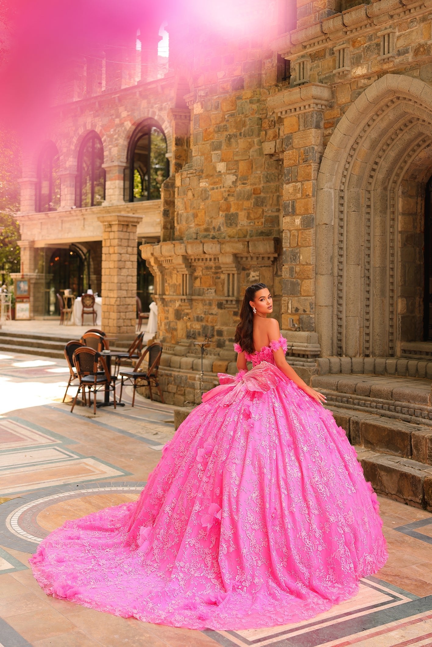 A person in a voluminous, bright pink ball gown poses in front of an ornate stone building with arched windows.