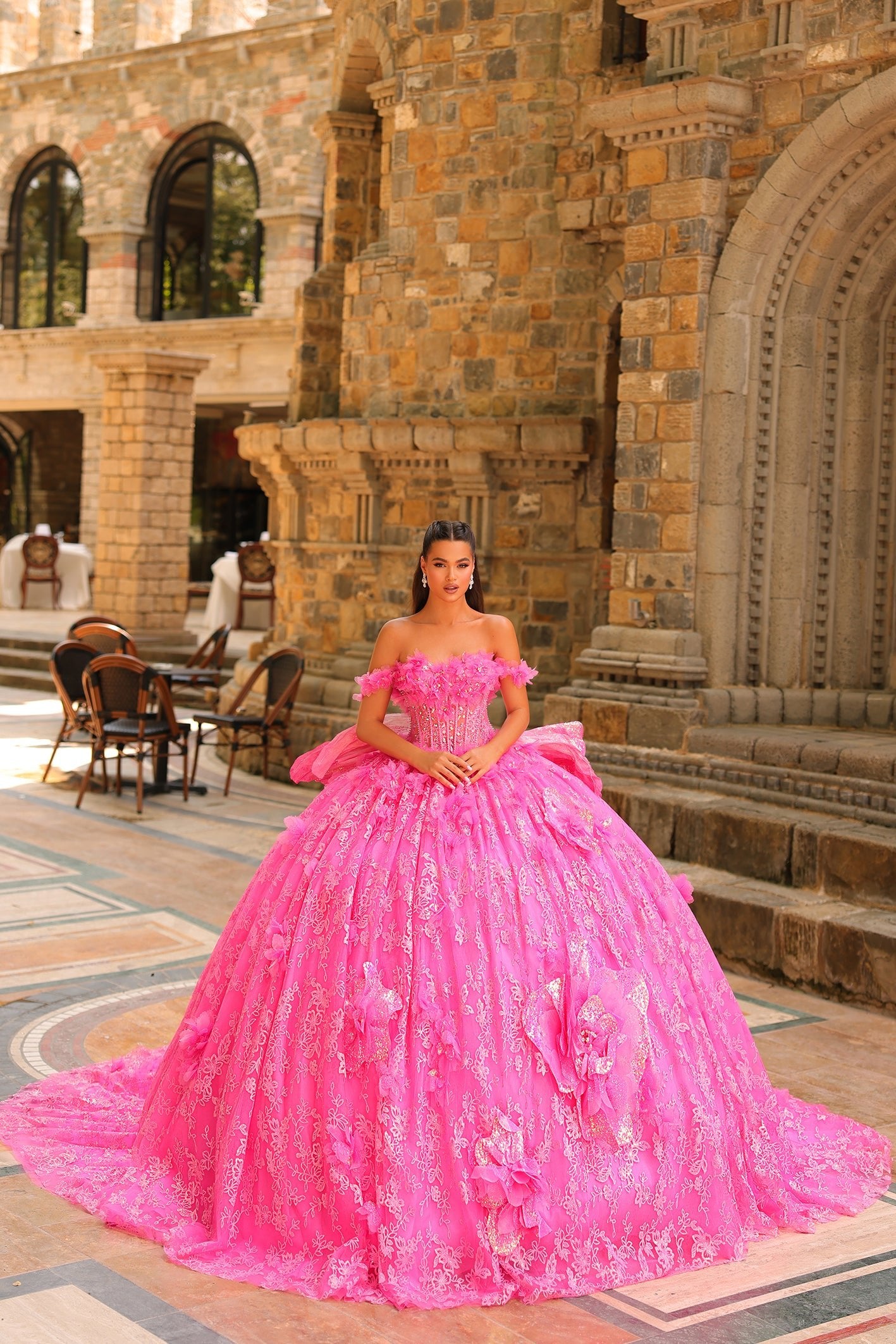 A woman wears a voluminous fuchsia off-shoulder ball gown with 3D floral details in a stone castle setting.