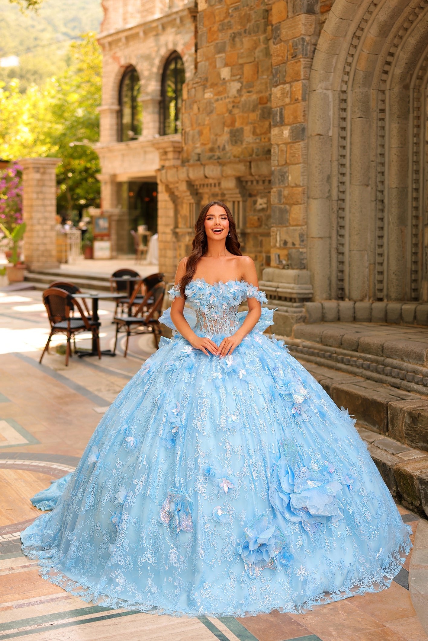 A woman in a light blue, off-shoulder ball gown with 3D floral details stands before a stone building.