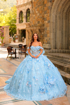 A woman in a light blue, off-shoulder ball gown with 3D floral details stands before a stone building.