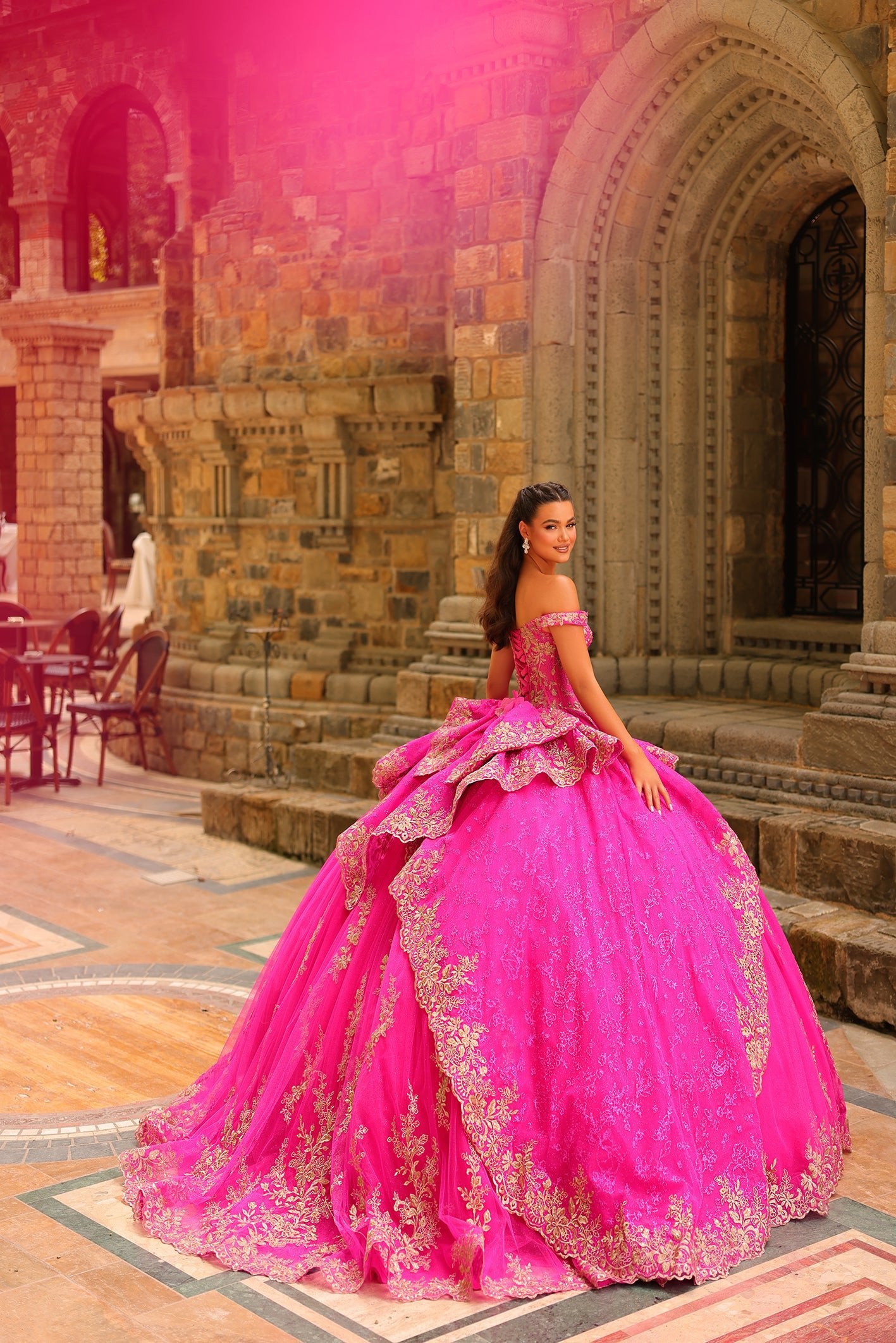 A woman in a vibrant pink ballgown with gold embroidery poses elegantly in a stone archway.