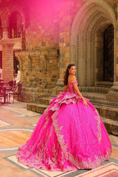 A woman in a vibrant pink ballgown with gold embroidery poses elegantly in a stone archway.