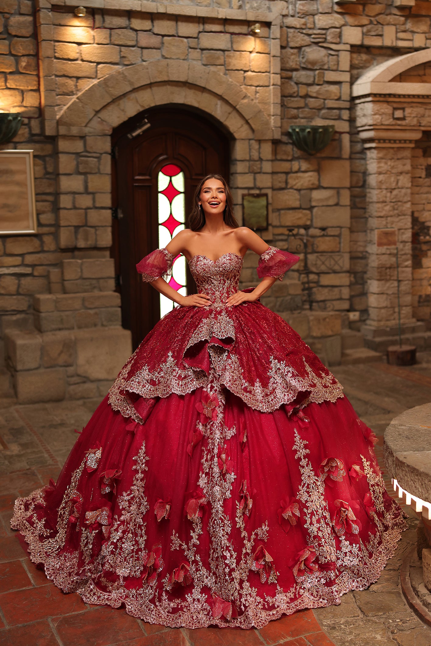 A woman wears a dramatic wine-colored ball gown with silver lace detailing in a stone castle setting.