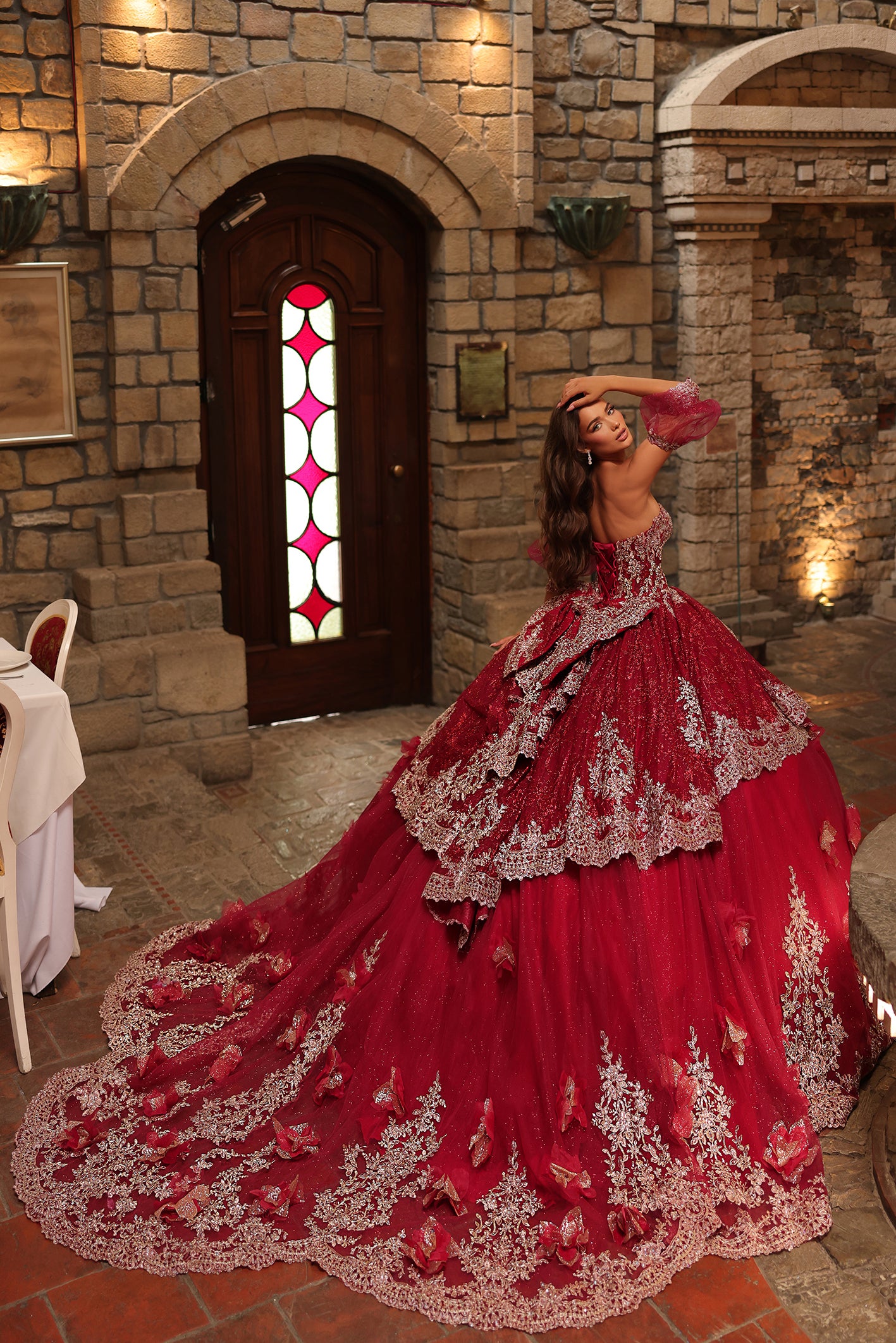 A woman in a dramatic red ballgown with silver lace stands in a stone castle with a stained glass door.