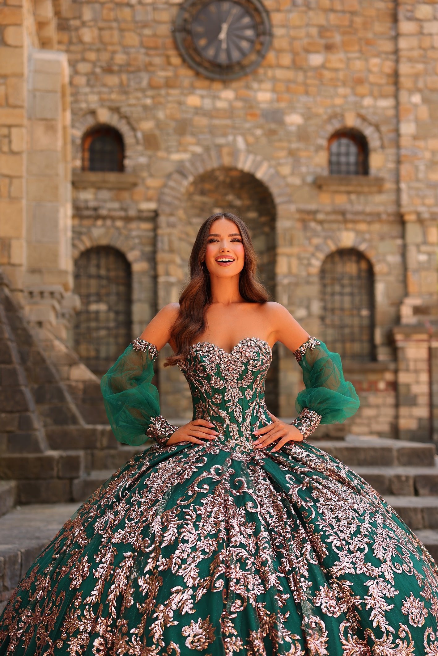 A woman in an emerald green and silver quinceañera gown poses dramatically in front of a stone castle.
