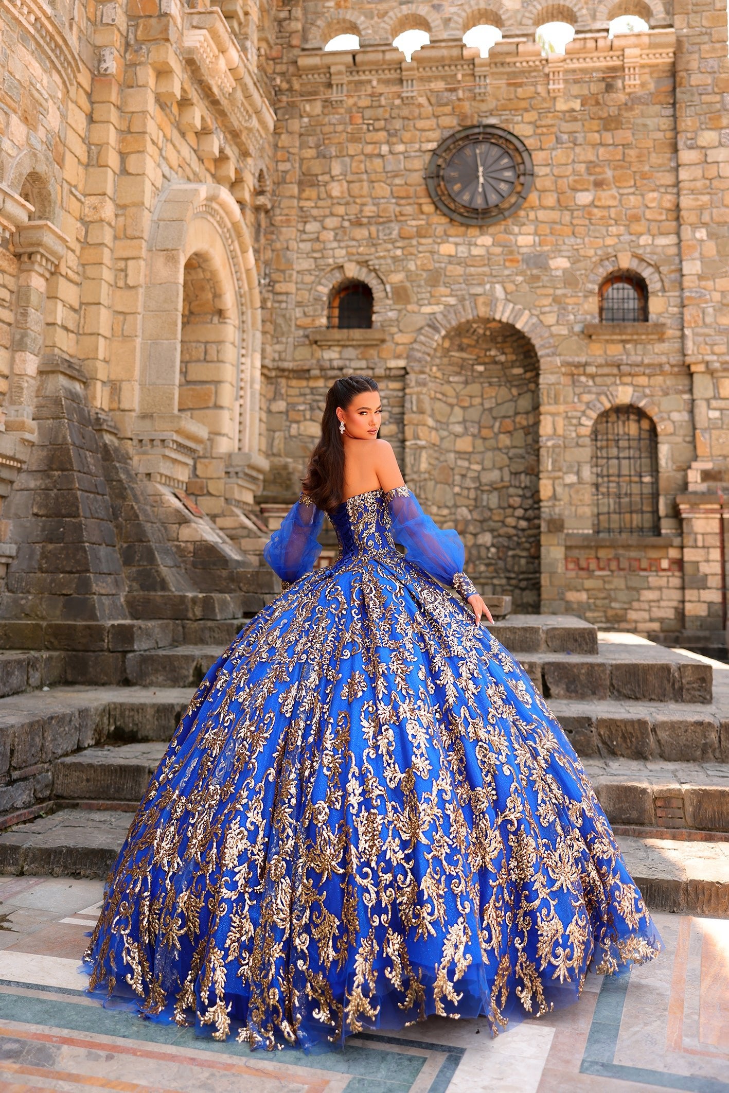 A woman wears an ornate blue and gold ball gown on stone steps with a medieval stone castle backdrop.