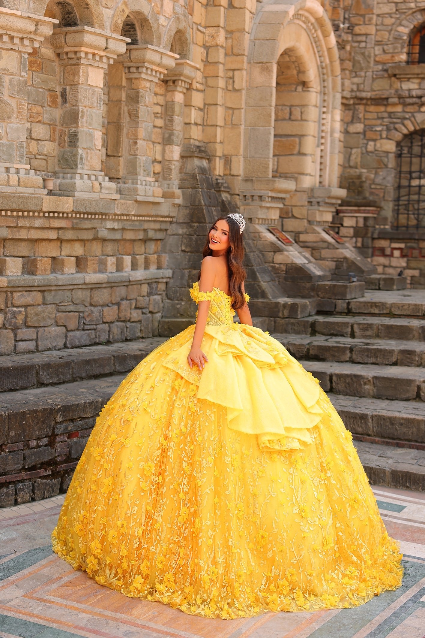 A woman in a voluminous yellow off-shoulder ball gown and tiara poses on stone steps with an architectural backdrop.
