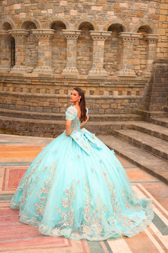 A woman in a teal ballgown with floral embellishments poses before a stone archway with intricate architecture.