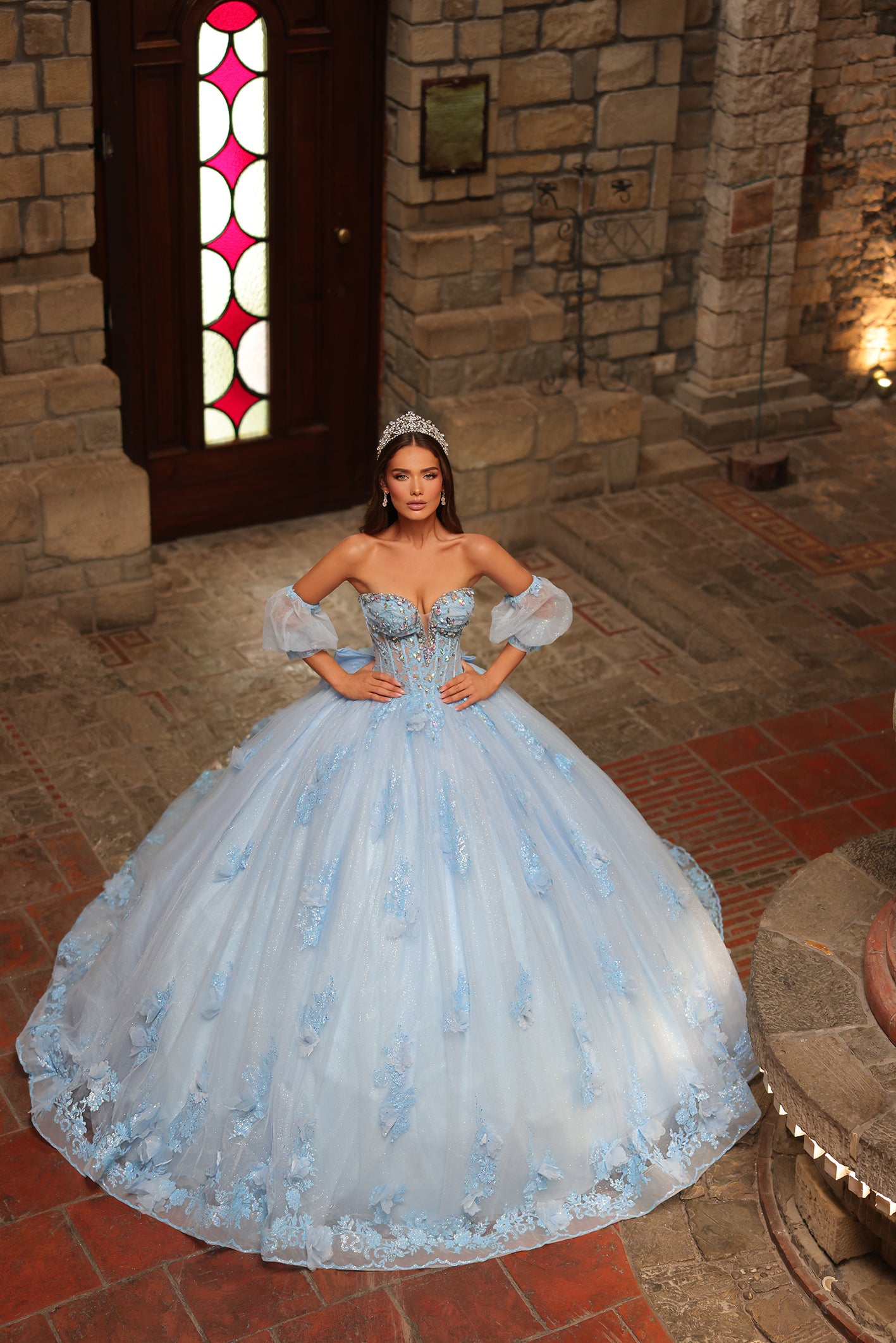 A woman wears a voluminous Light Blue ball gown with floral details, standing in a stone castle interior.