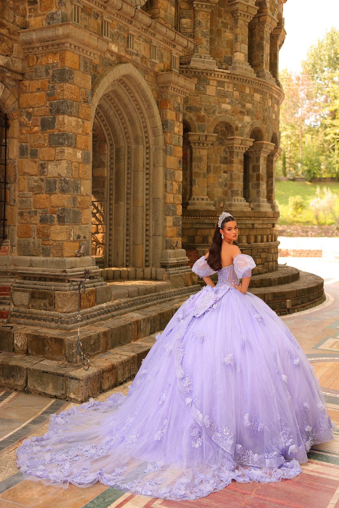 A woman in a lavender ball gown poses elegantly by a stone castle with ornate architectural details.