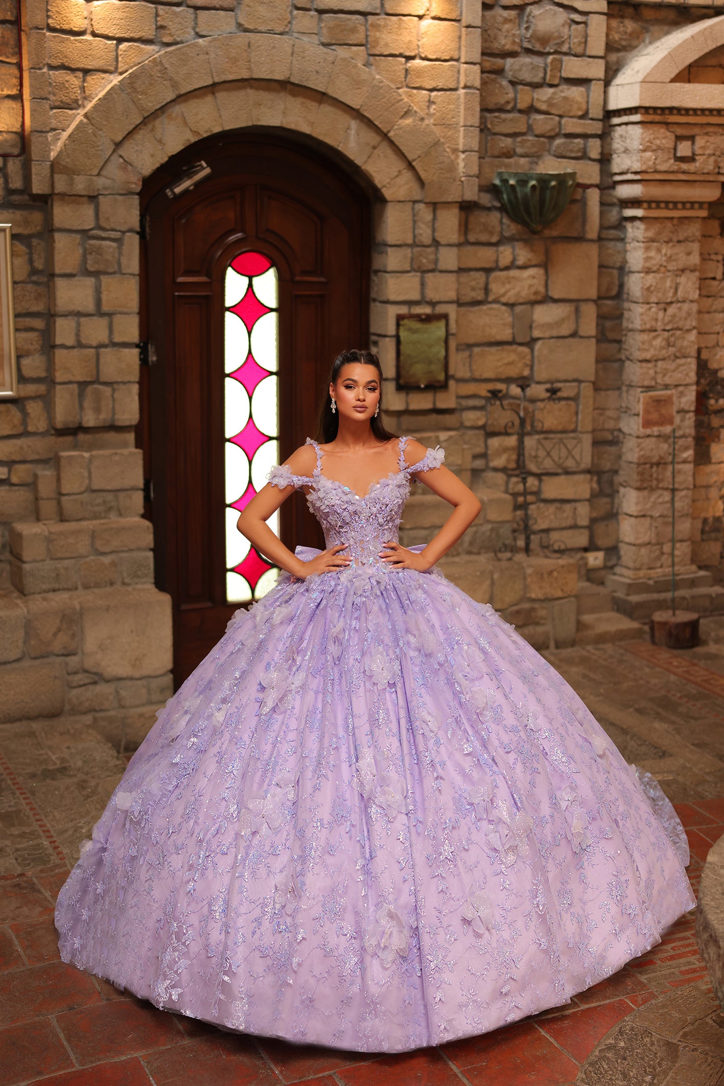 A woman wears a voluminous lilac ball gown with floral embellishments, standing in a stone-walled castle interior.