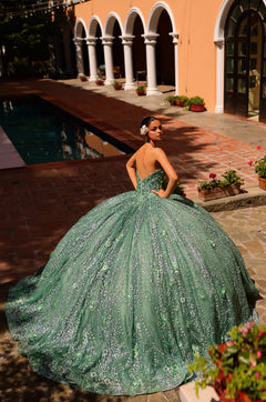 A person in an emerald green, sparkly ballgown poses by a pool near arched columns and potted plants.