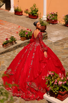 A woman in a vibrant red quinceañera gown with silver embroidery poses among flower-filled terracotta pots.