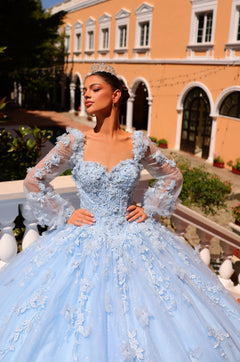 A woman in a light blue ball gown with floral embellishments stands elegantly before an orange building.