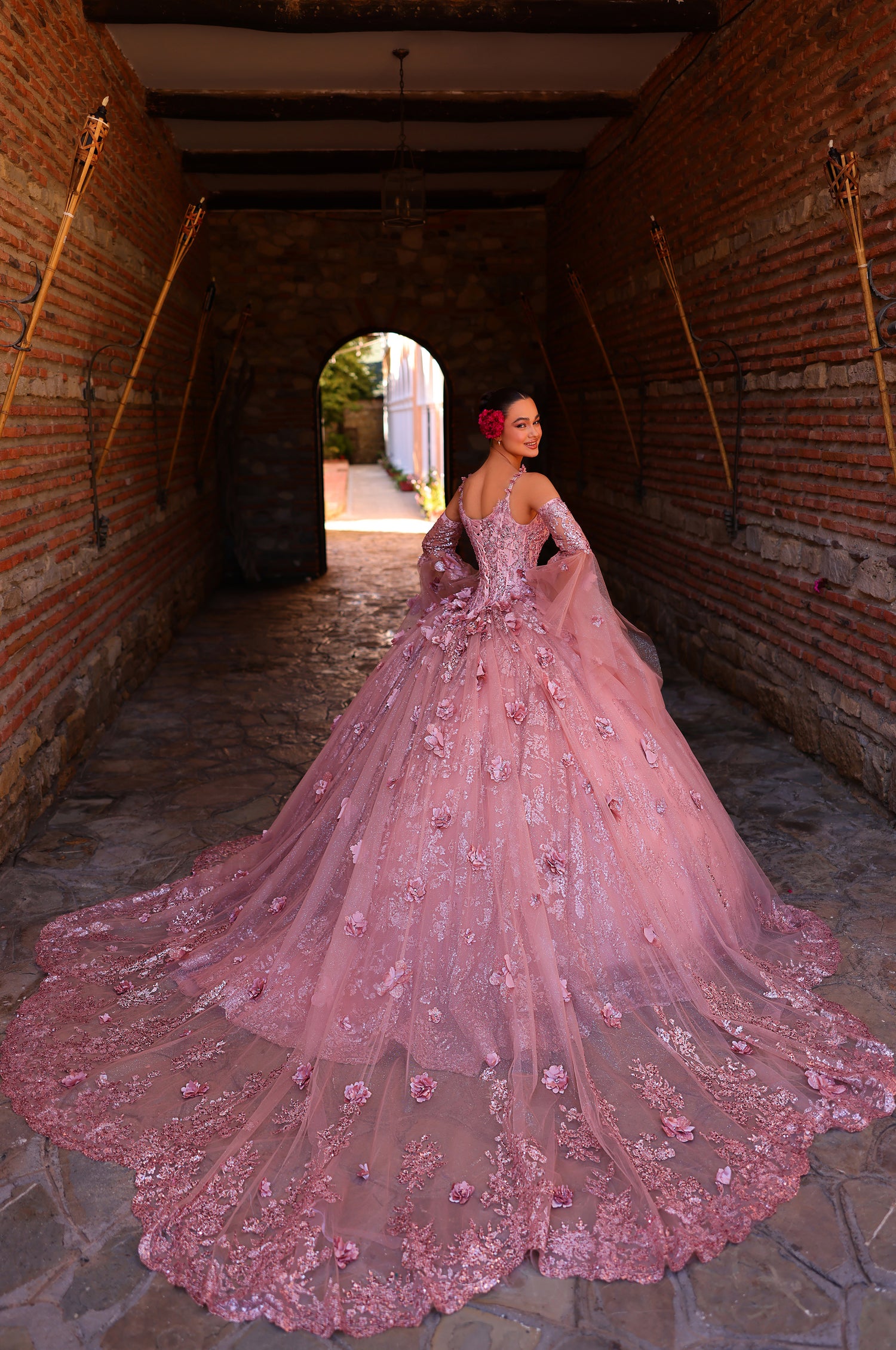 A person in a voluminous pink floral ball gown stands in a brick tunnel with an arched doorway.