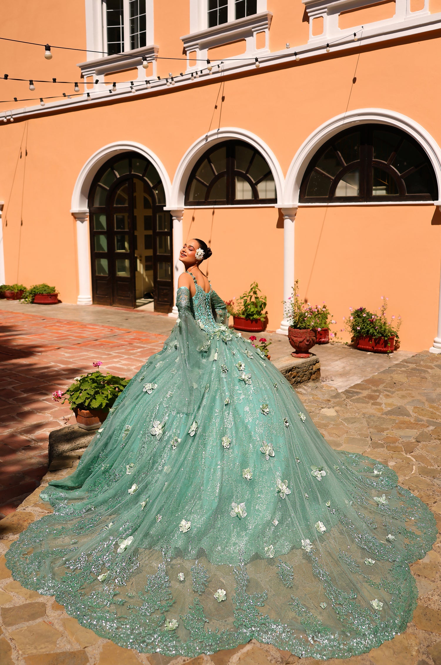 A person in a mint green, floor-length ballgown with 3D floral appliqués poses in an arched courtyard.