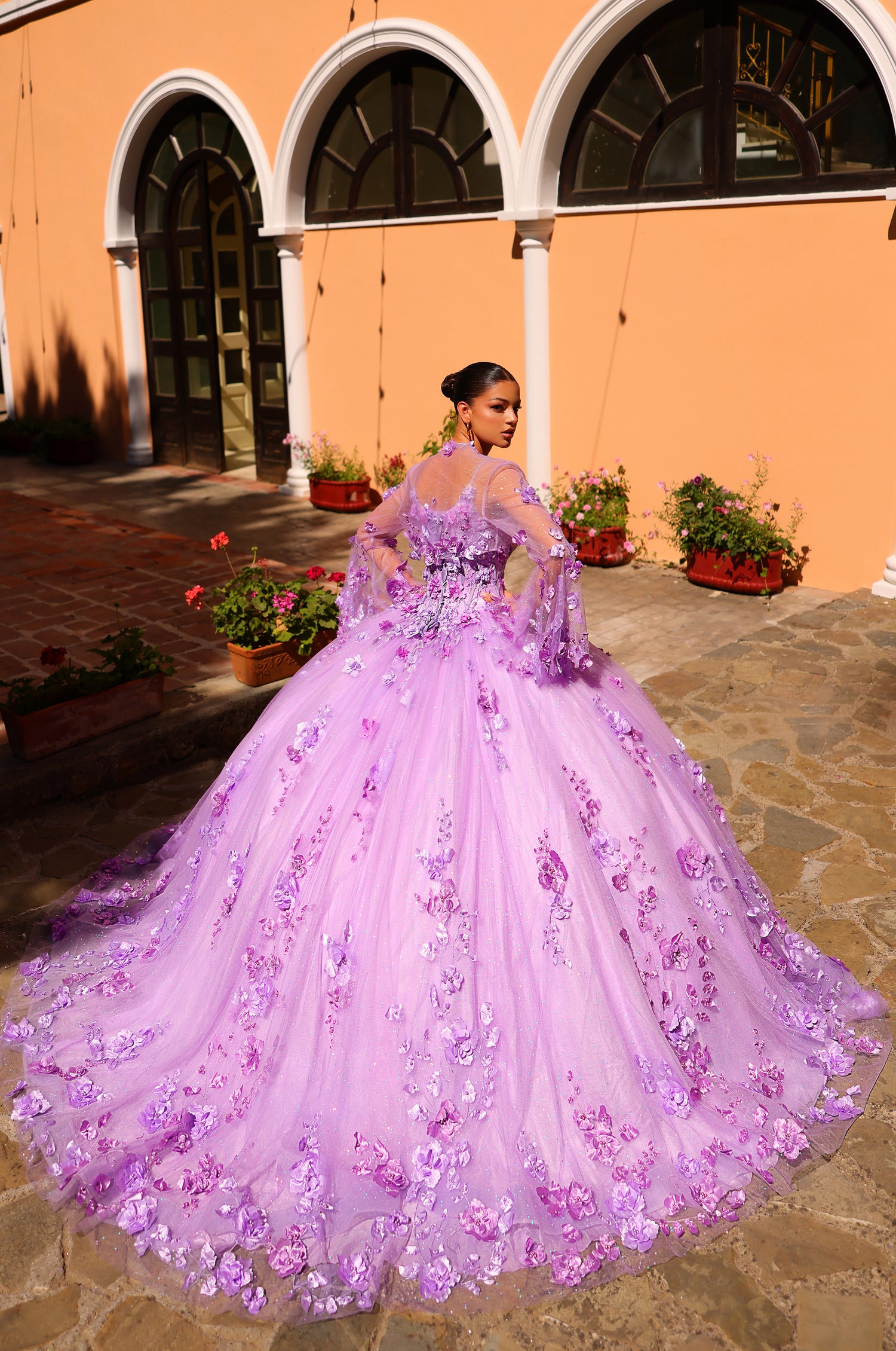 A lavender ball gown with 3D floral appliqués poses against an orange building with arched windows.