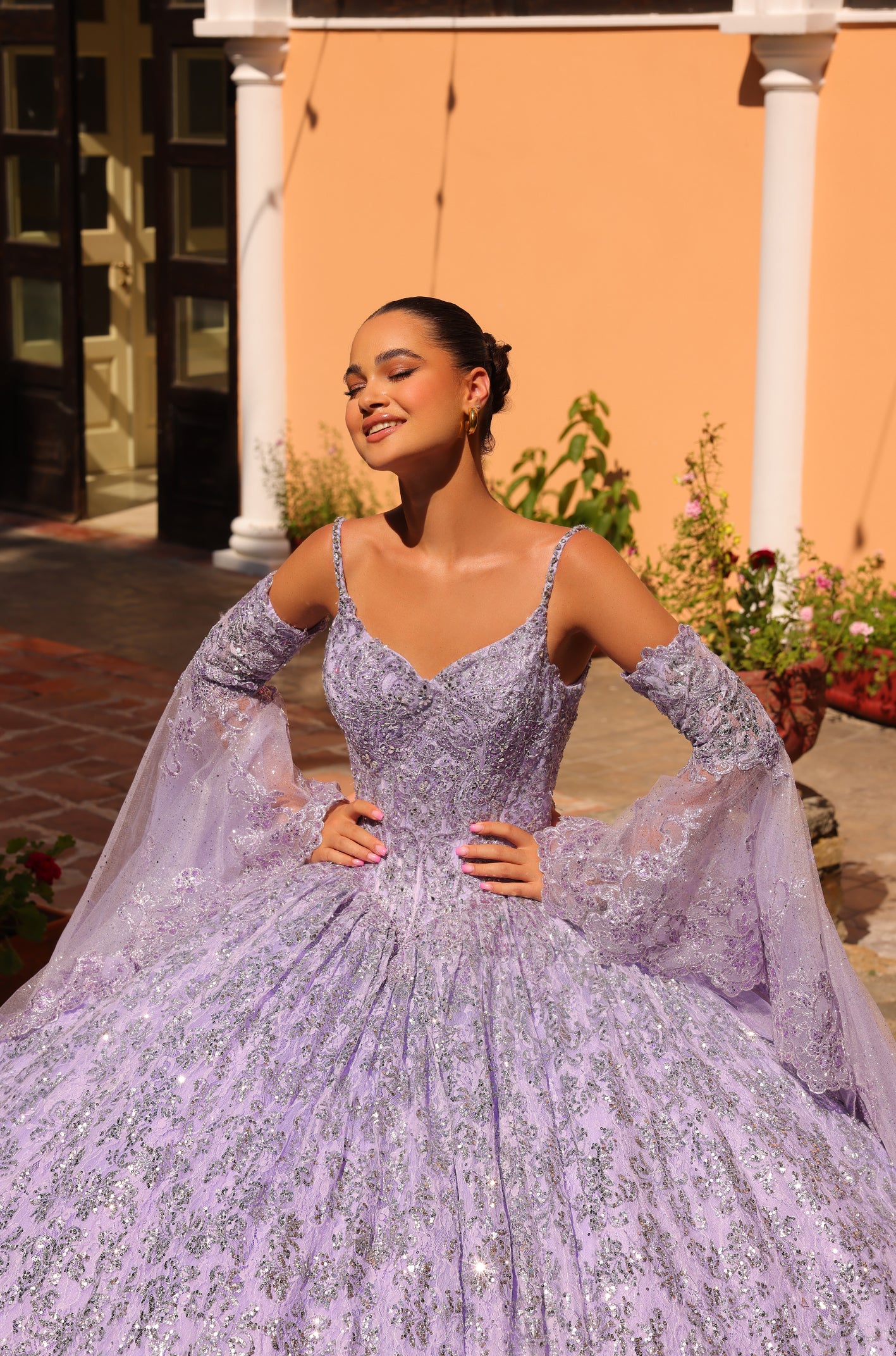 A woman in a sparkling lavender ball gown poses confidently in a sunlit courtyard with white columns.